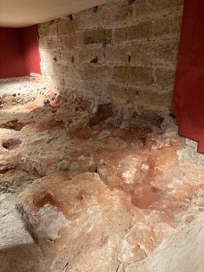 The image displays an excavated archaeological site in Cádiz, Spain, likely within a subterranean section of a historical structure such as the Roman Theatre of Gades or another urban excavation. The scene is an interior corner space with a discernible back wall, partial left and right walls, and an exposed ground level.

The dominant back wall is constructed from large, irregularly cut, light-colored stone blocks, arranged in horizontal courses with visible, aged mortar. Several small, rectangular apertures or niches are present in this stone wall. White, elongated mineral deposits or efflorescence cling to the lower portions of the stone wall, extending towards the ground. The left wall and a segment of the right wall are finished with a smooth, matte, dark red plaster or paint, which abruptly meets the rough stone and excavated ground. The ceiling, visible at the top, is a flat, light-colored surface.

The ground comprises an uneven, excavated layer of reddish-brown earth intermixed with lighter-colored, irregular stone fragments and debris. This substrate exhibits varying elevations, textures, and depressions, with the reddish hue being particularly pronounced in several areas. No individuals, modern objects, or specific actions are visible in the scene. The area is illuminated by an artificial light source, casting light from the upper left and accentuating the textures of the stone and earth.
