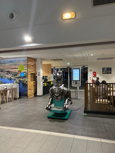 Inside a McDonald's restaurant, a silver horse ride stands near a digital ordering kiosk. The restaurant's interior includes light-colored walls, gray tiled floors, and recessed lighting. Behind the horse, there's a wooden slat wall with a landscape mural. A seating area with white chairs and tables is visible in the foreground. In the background, other patrons are sitting in a raised booth. A fire extinguisher and signage are on the wall behind the booth.