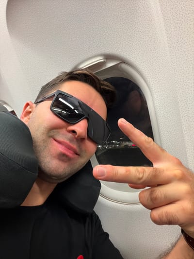 A man wearing sunglasses and a black shirt sits in an airplane seat with a headrest. He makes a peace sign with his hand, which is positioned near the plane window. The window's reflection shows city lights. The plane's ceiling is visible with its patterned surface.