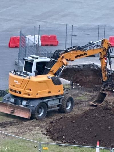 An orange and white Liebherr 914 compact excavator is parked on a dirt construction site in Reykjavik, Iceland. The excavator is positioned facing left, with its front blade lowered to the ground. A pile of dark soil is to the right of the excavator, and the excavator's bucket is digging into the soil. Red traffic barriers are visible in the background, along with a chain-link fence. The ground is covered with loose dirt and gravel. The sky is overcast.