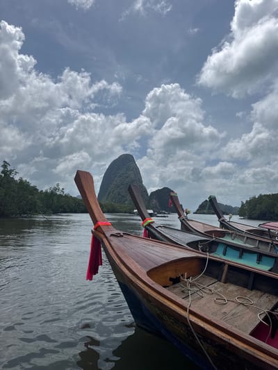 The image depicts a view of the water and several wooden longtail boats docked in the city of Ban Hin Rom, Thailand. In the foreground, the bow of a longtail boat is prominently featured, adorned with a vibrant pink cloth. Behind it, other longtail boats are lined up, each with decorative ribbons tied to their prows. The water is calm, reflecting the overcast sky and the surrounding lush green trees that line the shore. In the distance, a large, distinctive karst limestone formation rises majestically from the sea. The sky is filled with dramatic clouds, suggesting a potentially approaching storm or a day with dynamic weather. The scene conveys a sense of tranquility and the typical scenery of Thailand's coastal regions.
