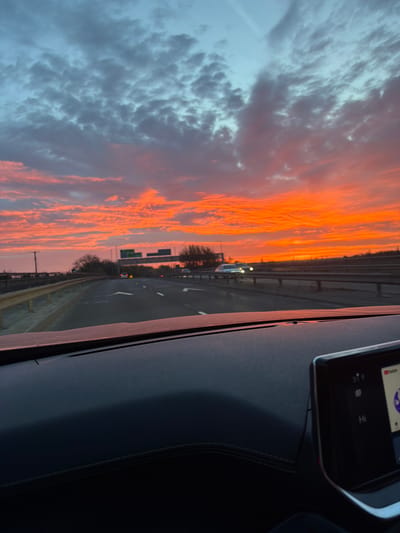 The image depicts a view from inside a car driving on a highway, likely in Garden City, United Kingdom, given the request. The highway is visible through the windshield with multiple lanes and guardrails. The sky is filled with a dramatic sunset, with streaks of orange and grey clouds. Part of the car's dashboard is visible, including a screen displaying a user interface and a portion of the seating area. There are no people visible. The action is the movement of the car on the highway. The interaction is the driver observing the sunset while driving. The environment is a combination of urban highway infrastructure and a dramatic sky.