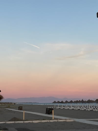The image depicts a serene beach scene at sunset in Pistula, Montenegro. The sky displays a gradient of colors, transitioning from a soft blue at the top to warm pink and orange hues near the horizon. A faint airplane contrail streaks across the upper portion of the sky. In the foreground, dark sand covers the beach, with a pathway made of concrete or paving stones. Along the pathway, there are two wooden structures, possibly serving as planters or bins. A small pile of sand is visible near the pathway. Further back, a row of thatched umbrellas stands over empty lounge chairs arranged on the sand, facing the calm sea. In the distance, a line of mountains creates a silhouette against the colorful sky, and a few indistinct figures can be seen walking near the water's edge. The overall impression is one of tranquility and the quiet beauty of a coastal evening.