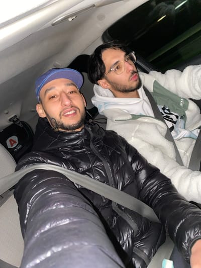 Two young men are sitting in a car. One is wearing glasses and a white jacket. The other man is wearing a blue cap and a black puffy jacket with the seatbelt on. The picture appears to be taken from the front passenger side, looking towards the back seat, where the two men are seated.