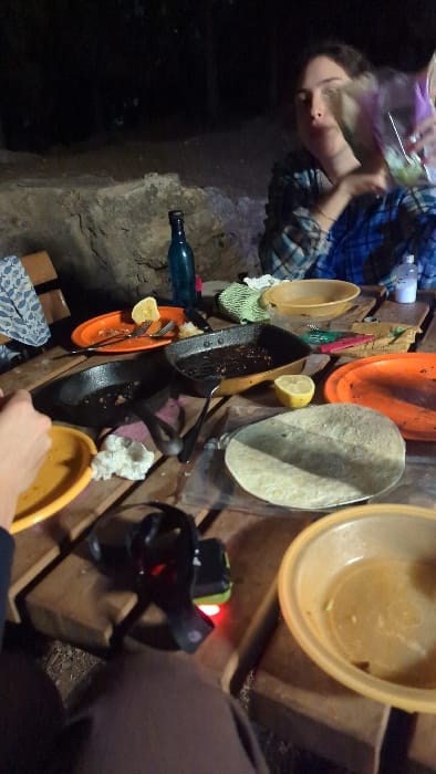 The image displays a nocturnal outdoor meal scene in Modiin-Maccabim-Reut, Israel. A wooden picnic table is central, laden with items from a recently consumed meal.
Two individuals are partially visible. On the right, a female subject with dark hair, wearing a blue and white plaid shirt, holds a clear plastic bag containing leafy greens; her face is partially obscured by motion blur. In the lower-left foreground, a hand holds an empty yellow plastic plate, belonging to an unseen individual.
Objects on the table include two dark frying pans with food residue, two orange plastic plates containing remnants (one with a corn cob piece, a fork, and a knife), an orange plastic bowl with liquid residue, a white tortilla on a clear plastic sheet, a blue glass bottle, two half-lemons, a green textured cloth, a small white plastic bottle, and a pile of white foodstuff. A black headlamp, exhibiting a red indicator light, rests on the table.
The background reveals a dimly lit, rough rock formation, confirming a natural, outdoor environment at night. The ground beneath the table appears to be dark earth.