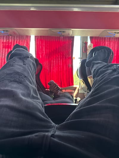 The image shows a view from inside a bus or other passenger vehicle, potentially near Izvorovo, Bulgaria. The focus is on a person's legs wearing dark, possibly ripped jeans, seated in the foreground. A second person is visible in the background, seated by a window with red curtains. The window shows a glimpse of the outside world. Interior elements of the bus or vehicle, such as the seat, the window frame, and possibly air conditioning vents are also visible.
