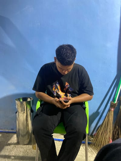 An image, originally rotated 90 degrees counter-clockwise, depicts an individual absorbed in a smartphone in what appears to be an indoor or covered outdoor setting in Gampongaceh, Indonesia.

The primary subject is a person, likely male, with short dark hair, seen from an elevated perspective looking down. They are seated on a lime-green chair, wearing a black short-sleeved t-shirt with a prominent, colorful graphic design on the chest, and black pants. Both hands are holding a dark-colored smartphone, indicating they are actively using it.

The background features a distinct, textured blue wall, possibly painted concrete. To the right of the subject, several brooms made of natural fibers/straw are leaning against the wall or in a corner, casting a shadow. Adjacent to the person's right leg and the chair, a shiny, metallic cylindrical object, potentially a trash can or part of a water dispenser, is visible. The floor beneath the chair and metallic object is a grey concrete surface.

The activity shows the person focused on their device, suggesting they are browsing, texting, or playing a game. The lighting, with distinct shadows, hints at artificial illumination or a specific time of day like evening or night. No strong emotions are discernible on the subject's face, only concentration. There is no visible text in the image.