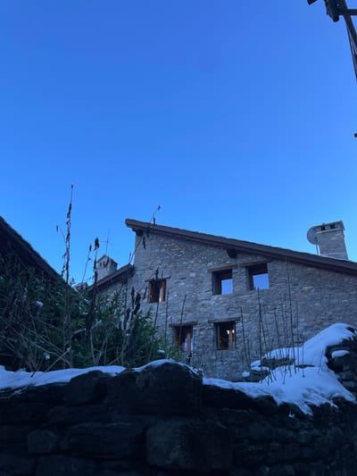 The photograph, when viewed in its intended landscape orientation (rotated 90 degrees clockwise from the uploaded version), depicts a traditional stone building under a clear, bright blue sky in a snowy, winter setting in Courmayeur, Italy.

The main subject is a rustic, multi-story building constructed from rough, irregular grey and brown stones, featuring a gabled roof made of dark material. A stone chimney extends from the roofline, and a small, partially visible satellite dish is mounted near it. Several rectangular windows are visible on the facade; some appear as empty frames or reveal dark interiors, suggesting the building might be unoccupied or seen from a distance.

In the immediate foreground, along the right side and bottom of the frame, is a rugged stone wall thickly covered with white snow. Scattered bare, dark tree branches and some evergreen bushes, also lightly dusted with snow, fill the lower and mid-ground, partially obscuring the base of the building.

The sky is a vibrant, cloudless blue, indicating a clear and sunny daytime. The presence of abundant snow and bare branches signifies cold winter conditions. There are no people or explicit activities taking place, contributing to a tranquil and serene atmosphere. No text is visible within the image. The scene captures the quiet beauty of an alpine winter day.
