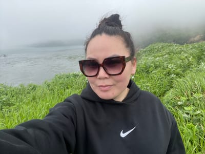 A woman is taking a selfie on a foggy day in Tikhoe, Russia. She is wearing a black Nike hoodie and large, tortoiseshell sunglasses. Her dark hair is pulled up into a bun. Behind her, the scene is dominated by lush green grass, with a body of water and a distant, fog-shrouded landmass visible in the background.