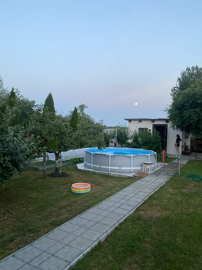 The image depicts a backyard scene in the village of Znamenski Selski Sovet, Belarus, during twilight. A large, circular above-ground swimming pool with blue water is the central feature, surrounded by a paved path. To the left of the pool, a small, inflatable ring pool with colored sections is visible on the grass. A pathway of grey concrete pavers leads from the foreground towards the pool and continues to the right, where a white building with a dark doorway is located. A woman in a black top and denim shorts stands near the doorway of the building, facing away from the camera. A hose is visible on the grass near the woman. The yard is filled with lush green grass and mature trees, including what appear to be fruit trees with some red fruit visible. In the upper right quadrant of the sky, a faint full moon is visible against a pale blue sky. The overall atmosphere is tranquil and residential.