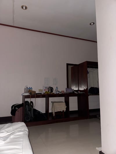 The image captures an interior room in Khorn Neua, Laos. The room features white walls and a light-colored tiled floor, reflecting ambient light. The ceiling is white, with two silver-rimmed recessed spotlights; a vertical discoloration or stain is visible near the left spotlight. A dark brown molding runs horizontally along the upper portion of the walls.

A long, dark wooden console table or desk is positioned against the back wall. On the table, from left to right, are plastic bags, several plastic water bottles, a woven bag, a white power strip plugged into an electrical outlet, an electric kettle, and additional water bottles and miscellaneous small items. Taped to the wall above the power strip are two sheets of paper with text, and a red sign displaying "FREE WIFI" and the number "59019".

Beneath the console table, a pink hardshell suitcase with a black garment atop it is visible on the left. Adjacent to it is a folded dark-colored baby stroller. A light-colored stool, draped with a white cloth, is centered under the table.

To the right of the console table, a dark wooden wardrobe or cabinet stands. The left door of the wardrobe is open, revealing a mirrored interior surface. The right section of the wardrobe is also open, showing a white t-shirt hanging on a hanger and a dark bag resting on the floor inside.

In the lower-left foreground, a portion of a bed with white sheets is visible. The room is unoccupied, and the overall lighting appears dim.