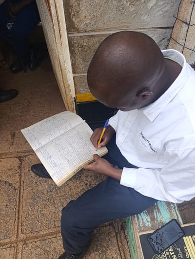 An overhead, slightly angled shot captures a man, likely a security guard, diligently writing in a large, open ledger in an outdoor or semi-outdoor setting in Kaplelwo, Kenya. The man is seen from above his head and shoulders, looking down at his task. He wears a white collared shirt with the text "INTERCITY SECURITY" emblazoned on the right chest, and dark trousers. He uses a yellow pencil with a blue tip to make handwritten entries in blue ink into the thick, aged notebook, which is filled with dense script.
He is seated on what appears to be a worn wooden bench or step with peeling light green paint. To his right, a black smartphone rests on the bench, face down or with a dark screen, partially obscuring an object with "ENTE" and "Book" visible on it. The ground beneath him is a textured concrete or paved surface embedded with small stones. In the background, to the left, another person's legs and feet are visible within a doorway or opening. A textured concrete block wall is seen to the right, featuring a yellow and black painted stripe near its base. The scene is brightly lit by natural light, suggesting it is daytime. The man appears focused and engaged in his record-keeping activity.