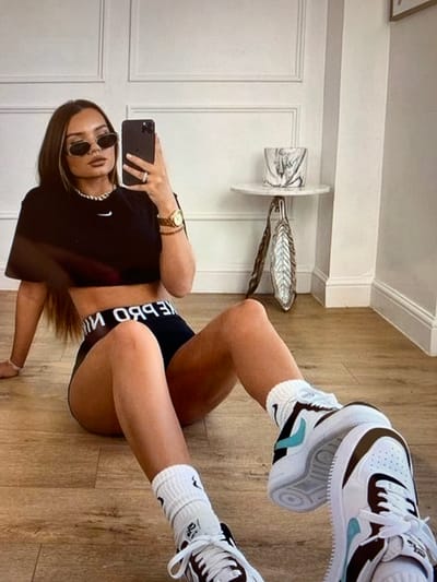 A young woman with long brown hair and sunglasses is sitting on a wooden floor, taking a selfie in her phone. She is wearing a black crop top, black shorts with "NIKE PRO" printed on the waistband, white socks, and white Nike sneakers with brown and teal accents. She is also wearing a necklace and a watch. In the background, there is a white wall with decorative paneling and a small round table with a decorative vase. The image appears to have been taken indoors, in a residential setting. The location is not specified in the image or surrounding context, therefore, it is not possible to determine if the location is in the city of Константиновка, Kyrgyzstan.