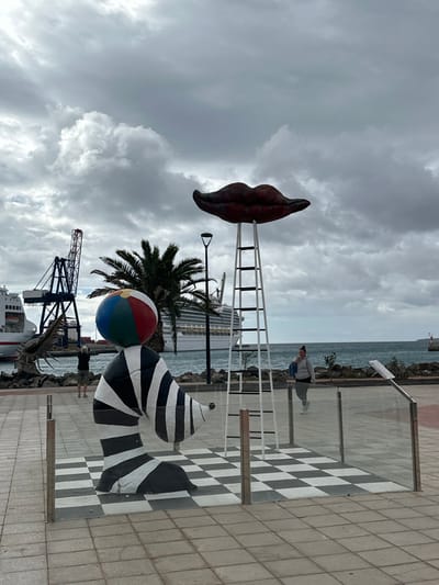 The image depicts a waterfront promenade in Puerto del Rosario, Spain, under an overcast sky filled with dense grey clouds. In the foreground, two art installations are visible. To the left, a large, stylized sculpture resembling a segmented, black and white striped creature, possibly a caterpillar or seal, has a multi-colored beach ball (red, blue, green, yellow, white segments) balanced on its head. This sculpture rests on a black and white checkerboard pattern on the paved ground. To the right, a large pair of reddish-brown lips is suspended atop a tall, white, ladder-like structure, also situated on a black and white checkerboard pattern. Both sculptures are partially enclosed by modern glass barriers with metal posts.
Behind the left sculpture, a person in a dark shirt and light shorts stands with their back to the camera, appearing to photograph or observe the harbor. Further left, part of a white and red ship and a prominent blue gantry crane are visible. A palm tree stands between the left sculpture and the background. In the mid-ground, a large white cruise ship is docked or moving in the ocean. To the right of the cruise ship, another person, dressed in a grey hoodie and dark pants, walks from right to left behind the glass barrier of the lips sculpture, carrying a blue bag on their shoulder. The background features the ocean with gentle waves, and a distant white lighthouse or beacon on a breakwater to the far right. The promenade itself is paved with light-colored square tiles.