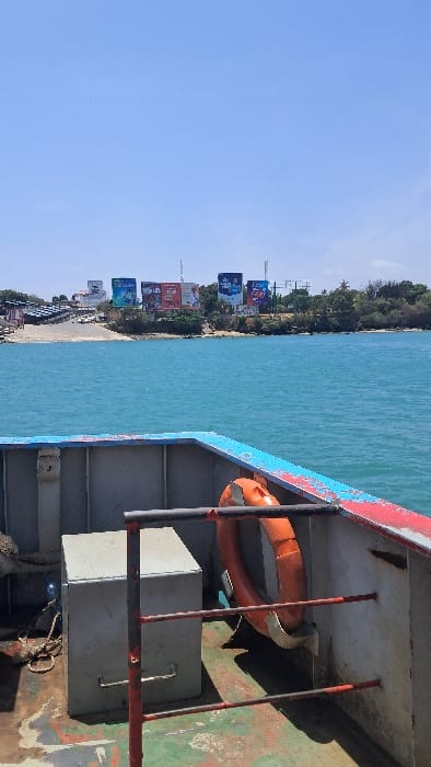 The image displays a view from the foredeck of a vessel, likely a ferry, positioned on a body of water in the Ganjoni/Shimanzi ward of Kenya, specifically within the Likoni Ferry channel.
In the foreground, the vessel's metal deck is visible, featuring worn blue-grey and red paint, exhibiting stains and peeling. An orange lifebuoy is mounted on the interior starboard bulkhead. A light-grey, rectangular metal storage box sits on the port side of the deck, near a coiled rope. A red and black metal railing runs horizontally across the mid-deck area.
The midground is dominated by clear, turquoise-blue water with minor surface ripples, indicative of calm conditions.
The background reveals a distant shoreline. A light-colored concrete ramp extends into the water, serving as a landing point for vehicles. Numerous vehicles are visible on and near this ramp, suggesting a queue or parking area. On the elevated land beyond the ramp, several large commercial billboards are prominently displayed. These billboards feature various advertisements, including one with "Safaricom" and "Binge more on the go" text. Green foliage, trees, and scattered communication or utility poles are present on the landmass. The sky above is clear and light blue, signifying fair weather. No individuals are explicitly visible in the scene.
