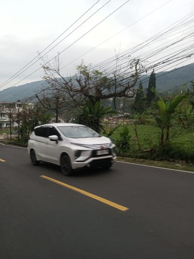 A blurry white Mitsubishi Xpander MPV travels on an asphalt road marked with a solid yellow line. The vehicle is moving past a landscape featuring grassy vegetation and trees, with a backdrop of a small mountain range under an overcast sky. Overhead, a dense network of power lines spans the scene. Behind the vehicle and slightly to the left, a multi-story white building is partially visible. Given the presence of the Mitsubishi Xpander, a model commonly used in Southeast Asia, and the landscape, the image is likely taken in a location similar to Taunggyi, Myanmar.