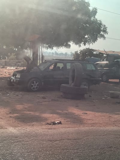 The image depicts an outdoor scene in Koton Karifi, Nigeria. A dark-colored station wagon or SUV, with its hood open, is positioned on a reddish-brown dirt surface. A plume of light grey smoke or steam rises from the engine compartment. Three used vehicle tires are stacked vertically next to the vehicle's rear passenger side. A second, partially visible dark-colored vehicle is parked behind it to the right. A large tree with dense foliage stands to the left rear of the primary vehicle, and several palm trees are visible in the distant background, along with low hills under a hazy sky. Structures, including one of cinder block construction and another with a corrugated roof, are present in the background. The foreground shows a darker, possibly paved strip of ground with scattered debris. No individuals are present in the frame.