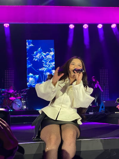 The image shows a female performer on a stage, likely during a concert or performance. She is holding a microphone and singing. She is wearing a white, long-sleeved shirt with wide sleeves, a black bottom, and fishnet stockings. Behind her, there is a large screen displaying blue and white flower graphics. Stage lighting creates a purple hue in the background. A drum set is visible at the bottom. Other people are present in the scene, including a musician and at least one person in the foreground holding a camera. The location is Salembaran, Indonesia.