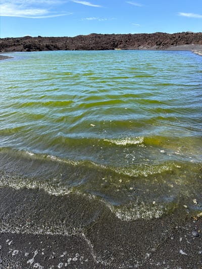 The image depicts a body of water, possibly a lagoon or inlet, bordering a dark, rocky shoreline under a partly cloudy, bright blue sky. The water has a greenish hue with visible wave patterns near the shore. The shoreline consists of a dark, volcanic-looking material, possibly black sand or lava rock, with some small patches of what appears to be foam or debris. The scene gives the impression of a coastal landscape, reminiscent of the volcanic terrain found in Yaiza, Spain.