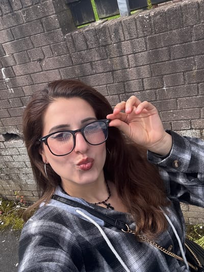 The image shows a young person, likely female, lying down on a paved surface. They are wearing glasses and a grey and black plaid shirt. A gold chain purse is slung over their shoulder. The person is holding their glasses with their right hand, and their lips are pursed. The background is a grey brick pathway with some moss and dry foliage. A dark wooden fence is visible on the left side of the image. The lighting suggests it is daytime, and the overall mood appears casual and playful. No visible text is present in the image. The setting appears to be outdoors, possibly in a park or garden area within Armagh, United Kingdom.