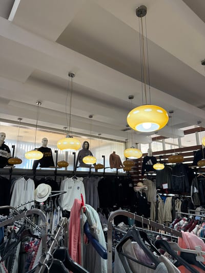 The image depicts the interior of a clothing store, likely a boutique or thrift shop, in the city of Acıpayam, Turkey. The store features rows of clothing displayed on racks, with mannequins placed behind them to showcase outfits. Overhead, several pendant lights with large, circular yellow shades and white polka-dot patterns hang from the ceiling, illuminating the merchandise. The ceiling itself is a tiered, white structure. The clothing visible includes a variety of garments such as hoodies, t-shirts, sweaters, and jackets, in an assortment of colors and styles. Some mannequins are dressed in branded sportswear, like the one with an Adidas logo on its shirt. The overall impression is one of a retail space organized for browsing and shopping.