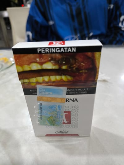 The image shows a close-up of a cigarette pack, likely a Gudang Garam Mild brand, placed on a white surface. The pack features a prominent warning label with an image of teeth and a red logo. There are also stamps and labels attached to the pack, including what appears to be a tax stamp with Indonesian currency denominations and the year 2020. In the background, a person wearing a blue jacket is partially visible, and some blurry yellow food items are on a plate to the left. The overall scene suggests an indoor setting, possibly a table in a home or a restaurant. There is no discernible indication of the city of Miruektaman, Indonesia in the image.