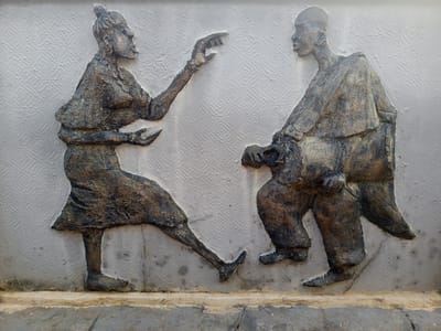This media file captures a wall relief sculpture depicting two human figures in an outdoor setting, likely in Osogbo, Nigeria. The sculpture is rendered in a dark, earthy, and textured material, possibly bronze or a similarly patinated metal, set against a light-colored wall with a distinct, square-patterned texture.
The figure on the left is a woman, portrayed in a dynamic pose. She is facing right, towards the male figure. Her left arm is raised and extended forward, her hand open as if gesturing or pointing with emphasis during a conversation. She appears to be wearing traditional clothing, including a skirt or wrapped lower garment, and her hair is styled in a bun or topknot.
The figure on the right is a man, depicted in a slightly crouched or seated posture, facing the woman. He wears flowing robes or draped clothing. His head is turned towards her, suggesting he is actively listening or observing. His hands are positioned at his waist, though the details of what he might be holding or doing with them are indistinct.
The interaction between the two figures suggests a lively dialogue, a negotiation, or perhaps a storytelling session, with the woman appearing to be the more animated speaker. The overall style of the relief reflects an African cultural context. The lighting suggests daylight, but there are no strong shadows to indicate a specific time of day or weather. No visible text is present in the image.