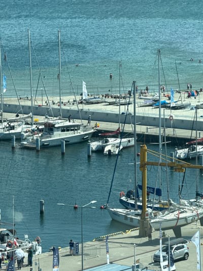 The image captures a bustling scene at a marina in Gdynia, Poland, likely during a summer day, given the presence of people enjoying the beach and water. In the foreground, several sailboats are moored, their masts reaching towards the sky. One prominent white motorboat, identified by the name "DRAGON" on its hull, is docked at the pier, with several individuals visible on its deck and near it. A silver car is parked on the dock, with flags and signs indicating a public event or area.

The middle ground of the image shows more sailboats and a concrete pier, with a sandy beach beyond. Numerous people are scattered across the beach, some sunbathing, others wading in the shallow water or engaged in activities like cycling. The turquoise water of the Baltic Sea forms the backdrop, with ripples indicating gentle waves. Several small boats and buoys are visible in the water.

The background reveals more of the coastline, with a distant view of buildings and infrastructure that are characteristic of a port city. The overall impression is one of a vibrant maritime location, combining leisure activities with the operational aspects of a marina. The sky appears clear and bright, suggesting fair weather.