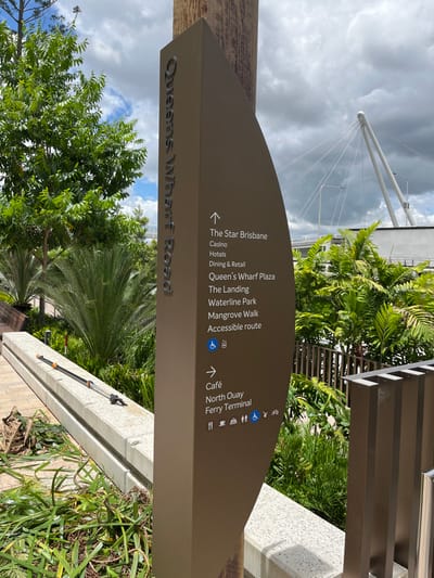 An outdoor shot captures a brown, angled directional sign pointing to various locations around Brisbane City, Australia. The sign, mounted on a wooden post, prominently features "Queens Wharf Road" in embossed lettering on its side.

The sign's main face lists destinations for pedestrians:
- **Forward/Right arrow:** "The Star Brisbane," "Casino," "Hotels," "Dining & Retail," "Queen's Wharf Plaza," "The Landing," "Waterline Park," "Mangrove Walk," and "Accessible route" (accompanied by a wheelchair icon).
- **Left arrow:** "Café," "North Quay" (with a bicycle icon), and "Ferry Terminal" (with a ferry icon and a food/drink icon).

The background shows a cloudy sky with patches of blue, suggesting a partly cloudy day. A white, cable-stayed structure, likely part of a bridge or modern building, is visible against the sky. Lush green foliage, including palm trees and other shrubs, surrounds the sign, indicating a well-landscaped urban or park environment. A grey concrete barrier runs along the right side of the frame, separating the pathway from the greenery. No people or activities are visible, emphasizing the informative nature of the signage for navigation within the Queen's Wharf precinct.