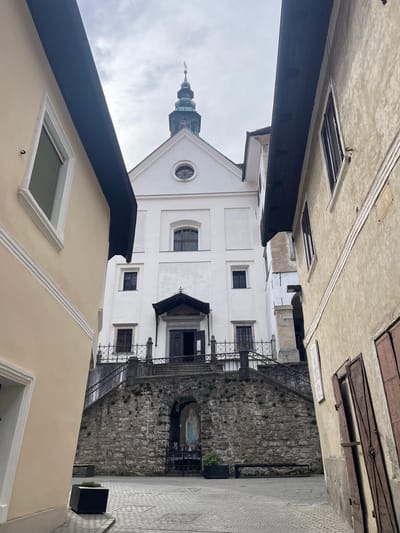 The image shows a narrow street view in Skofja Loka, Slovenia, with a church dominating the center. The church is white with a dark spire, featuring windows and a central entrance with stairs leading up. Walls of buildings line both sides of the street. The buildings are different colors: one is white, and the other is beige. Shutters can be seen on one of the buildings. On the right side, a stone wall with metal railings and stairs, along with a religious shrine, is visible. The sky is overcast.