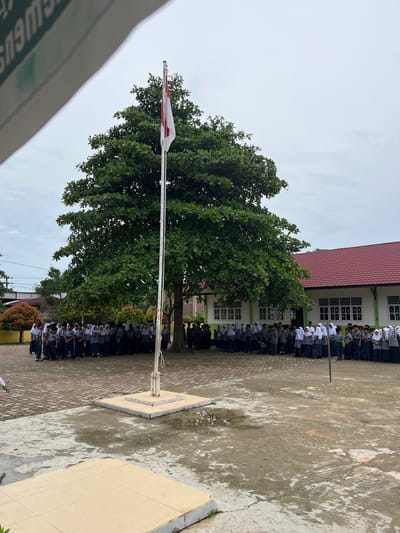 A group of students, some wearing headscarves, stand in formation on a paved outdoor area in front of a building. The students are dressed in school uniforms, consisting of dark pants and light-colored shirts, with some wearing grey overshirts. A flag pole with a red and white flag is positioned in the center of the formation. A large tree with lush green leaves is located behind the students, casting shadows on the ground. The building in the background has a red tiled roof and white walls with multiple windows. The sky is overcast, suggesting a cloudy day. The area appears to be a school courtyard.