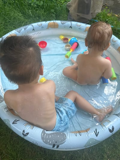 Two young children are playing in a shallow, inflatable paddling pool on a grassy lawn. One child, with light brown hair, is facing away from the camera, sitting with their legs extended in the water. They are naked. The other child, with dark brown hair, is in the foreground, facing the camera and looking to their right. They are wearing blue patterned swim shorts. Both children are wet and have water droplets on their skin.
Several brightly colored toys are scattered in the pool, including a yellow duck, a red ball, and a toy water squirter with pink, blue, and green components. The paddling pool itself is decorated with images of whales, rainbows, and other playful patterns. The water is clear and reflects the sky.
The scene appears to be outdoors on a sunny day, likely in the afternoon, given the lighting. The presence of the paddling pool and the children's attire suggest it is a warm day suitable for water play. The background shows green grass and some indistinct objects, possibly garden furniture or other outdoor items. The overall impression is one of a casual, summery activity. There is no visible text in the image.