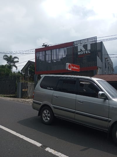 A silver minivan travels along a paved road next to a gray building with a black and red sign reading “KT HOTEL.” A smaller red and white sign with “RedDoorz” is displayed below. Across the road, behind a black fence, is a small house with a gable roof, partially obscured by trees. The sky is overcast.
