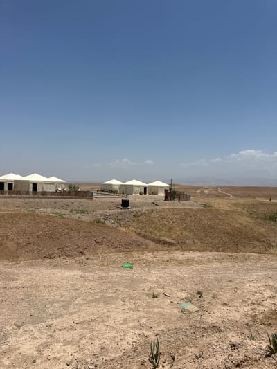The image depicts a series of white tents in a desert-like environment, possibly a luxury campsite, under a clear blue sky. The terrain is arid and consists of dirt and sparse vegetation. In the background, there are hills or mountains under a hazy sky. The location is near Sidi Badhaj, Morocco. There are no people visible. Some tents have visible openings, suggesting they are inhabitable. The image is rotated 90 degrees counterclockwise. A few agave plants are visible in the foreground. There are green and blue objects on the ground.