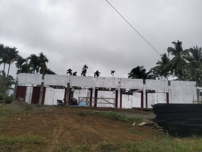 The image depicts an outdoor construction site under a cloudy sky. In the background, a partially constructed building with white walls and visible structural elements is present. Red corrugated metal panels are installed in some of the openings. Several palm trees are visible behind and above the structure. In the foreground, a pile of dark metal bars is neatly stacked on the right side of the image. A motorcycle is parked near the building, and a mound of dirt is situated to the left. The ground is covered with rough terrain, dirt, and some patches of green grass. The overall scene suggests ongoing construction in a tropical or subtropical location.