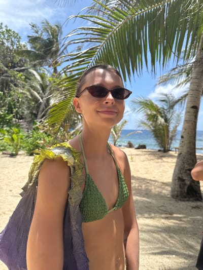 A woman in sunglasses and a green bikini stands on a sandy beach, likely in a tropical setting. Palm trees frame the scene with the ocean visible in the background. The exact location in Santa Monica, Philippines is undetermined from the image.