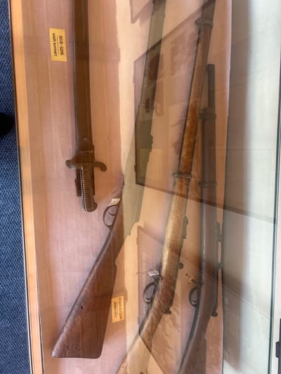 This image depicts a display case containing several antique firearms, likely in a museum or historical exhibit. The firearms are arranged vertically and appear to be rifles with bayonets attached to at least one of them. One rifle is partially visible on the left, showing its wooden stock and the bayonet mounted on the barrel. To the right, two more rifles are visible, their wooden stocks and metal components clearly discernible.
A yellow label is attached to the wooden stock of the rifle on the left, with text that appears to be in Cyrillic script and the English words "KNIFE BAYONET". Another yellow label is present on the wooden stock of the rifle on the far right, with Cyrillic script and the English words "BERDAN RIFLE". The background is a textured, light-colored surface. The glass of the display case creates reflections, making some details appear layered or slightly obscured. The context provided indicates this display is located in the city of Belogradchik, Bulgaria.
