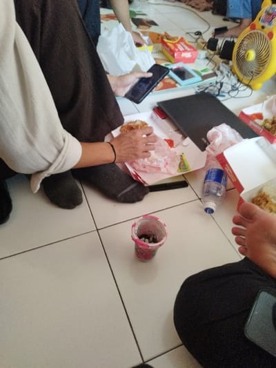 An indoor scene in Muka, Indonesia, depicts a casual gathering with individuals seated on a light-colored tiled floor with a patterned mat in the background. In the foreground, a person wearing dark brown trousers and a light-colored, long-sleeved shirt with rolled cuffs is holding a black smartphone in their left hand while their right hand holds a partially unwrapped fried chicken sandwich or burger over an open white fast-food box. A black wristband is on their right wrist, and their feet are clad in dark grey socks. To their right, the bare foot and lower leg of another individual, wearing dark trousers, are visible, with a smartphone resting on their lap.
Various objects are scattered on the floor: an open red and white fast-food box containing fried chicken and rice, a clear plastic water bottle lying on its side, and a small transparent plastic cup with pink trim containing dark cylindrical objects, possibly cigarette butts or discarded food. A black laptop is open, and a dark pen rests nearby. In the background, a yellow electric fan shaped like a cartoon character is plugged in, surrounded by cables. Other fast-food packaging, including a red box for fries and a crumpled white plastic bag, are also visible. A light blue tablet or phone lies on the patterned mat. Partial views of other individuals' limbs suggest at least two more people are present in the background.