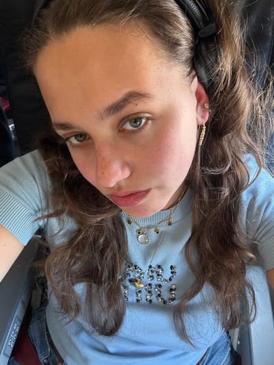 A young woman with brown hair is looking at the camera from a low angle. She is wearing a light blue sweater with beaded designs on the front and layered necklaces. She has multiple piercings in her ear and is wearing black headphones. The background appears to be an airplane interior, with a control panel visible in the lower right corner. The image does not contain sufficient information to infer the location is the city of Peschiera Borromeo, Italy.