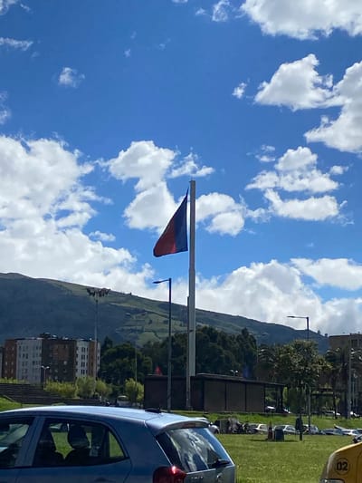 The image depicts a view in Quito, Ecuador, characterized by a blue sky dotted with cumulus clouds. A flag, colored blue and red, is visible on a pole in the mid-ground. In the background, a green hillside slopes down to a collection of buildings, suggesting an urbanized area. A car is partially visible on the right side of the image. Other cars can be seen parked on a grassy field to the right.