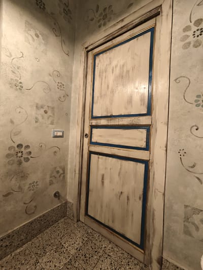 The image shows a closed, white, paneled door with distressed wood detailing and blue and gold trim. The door is set into a corner of a room with walls decorated with a faded floral stencil pattern. A light switch with three toggles is visible on the wall to the left of the door, and a small pipe or valve emerges from the wall near the floor. The floor is tiled with a speckled, terrazzo-like material. The lighting suggests an indoor environment, possibly a residential hallway or entryway. The location is identified as Pozzuoli, Italy. No people are visible in the image, and no specific actions are depicted.
