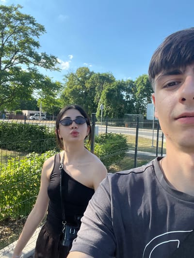 This media file is a selfie taken by two people outdoors on a sunny day. The subjects are a young man and a young woman. The man is in the foreground, with his face partially visible at the top of the frame. He has dark hair and is wearing a dark t-shirt. The woman is to the right of the man, and her face is more prominent. She has dark hair, is wearing sunglasses and a black tank top, and is making a pouty face.

The background shows a bright blue sky with some scattered clouds and green trees. There is a metal fence and some vegetation behind the subjects. In the distance, there appears to be a building and possibly a road or railway line. The lighting suggests it is daytime, likely midday or afternoon, given the bright sun and clear sky. The overall atmosphere appears casual and relaxed, consistent with an outdoor activity on a pleasant day. No text is visible in the image.