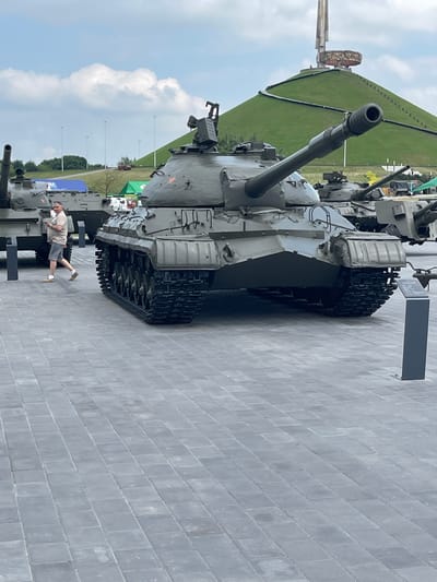 The image shows a large, grey tank displayed outdoors on a paved area. The tank is the primary subject, with its turret and tracks clearly visible. In the background, a grassy hill slopes upwards, topped by a monument. The sky is cloudy, suggesting it might be daytime with overcast weather. A person in shorts and a t-shirt is standing near the tank, appearing to observe it. To the left of the tank, the environment includes trees, poles with lights, and tents, indicating an outdoor exhibition or museum setting. The scene appears to be a memorial or a military museum due to the presence of the tank and monument. No specific text is clearly legible in the image.