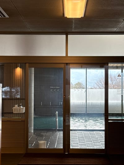 A brightly lit wall fixture casts light on a wall panel. Adjacent to the panel is a multi-pane window with a sliding door. The window offers a partial view of an outdoor area. Signs reading "Slide to Open" with arrows are on the glass. Beyond the glass, a tiled patio and water feature are visible. The scene's lighting and composition suggest an indoor-outdoor connection with a focus on functionality and design.