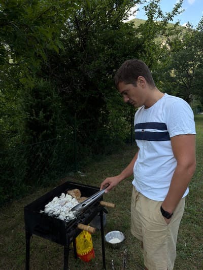 A young man wearing a white t-shirt with navy and red stripes and beige shorts stands to the right of a black charcoal grill. He is looking down at the grill, holding a pair of metal tongs over raw food that is being cooked. The grill is placed on a grassy area with trees and foliage in the background. To the left of the grill, a green chain-link fence encloses more vegetation. A yellow bag, likely charcoal, is hanging from the side of the grill. In front of the grill and to the right of the yellow bag, a small metal bowl containing a white powder is visible. The setting appears to be an outdoor location, possibly in or near Bistrica, Montenegro, given the hilly, forested environment. The sky is partially visible through the trees, with some blue sky and clouds.