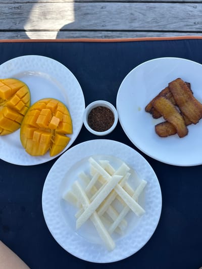 The scene is set outdoors in San Pedro Town, Belize, on a wooden deck, with a dark blue placemat featuring an orange border. Three white, round plates with a textured surface are arranged on the mat. The plate on the left holds two halves of a ripe mango, meticulously scored into cubes. The plate on the right contains four rectangular pieces of golden-brown fried plantain. The plate at the bottom presents a cluster of white, uniformly cut rectangular sticks, consistent with jicama or a similar root vegetable. A small white bowl, centrally positioned, contains a dark brown, granular substance, likely a spice blend. Natural sunlight casts distinct shadows on the wooden planks in the background. A light-skinned forearm is partially visible in the bottom-left corner of the frame, indicating the presence of a person.