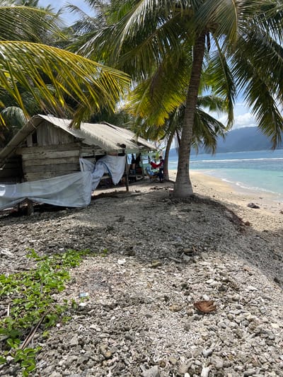 In Kruengtunong, Indonesia, an old, weathered wooden shack with a corrugated metal roof stands on a beach littered with coral fragments. A white tarp is draped over part of the structure. Coconut palms with lush green fronds overhang the scene, casting dappled shadows. Two individuals are visible near the shack; one is sitting in a folding chair, while another stands nearby, possibly interacting with a hammock. The turquoise ocean stretches out towards a distant, hazy coastline, with gentle waves lapping at the sandy shore. The sky is partly cloudy.