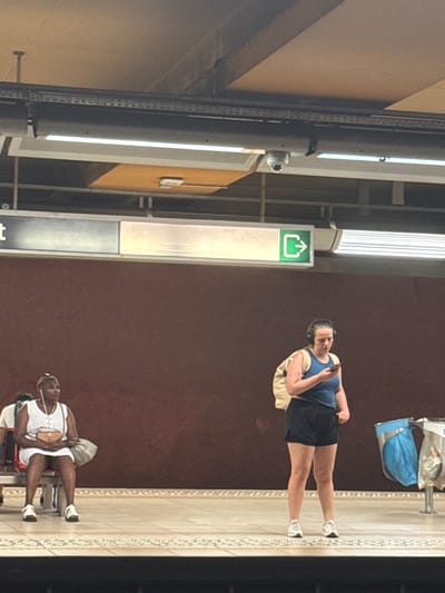 In the city of Molenbeek-Saint-Jean - Sint-Jans-Molenbeek, Belgium, at a subway station, two women are visible on the platform. On the left, a woman with dark skin and braided hair is seated on a bench, wearing a white dress, white shoes, and carrying a purse. She has headphones in her ears. On the right, a woman with light skin and dark hair, wearing headphones, is standing and looking at her phone. She is dressed in a blue tank top and black shorts and has a beige backpack on. The background features a dark brown wall, a ceiling with lighting and a security camera, and illuminated signs indicating an exit. To the right, a trash receptacle with blue bags is present.
