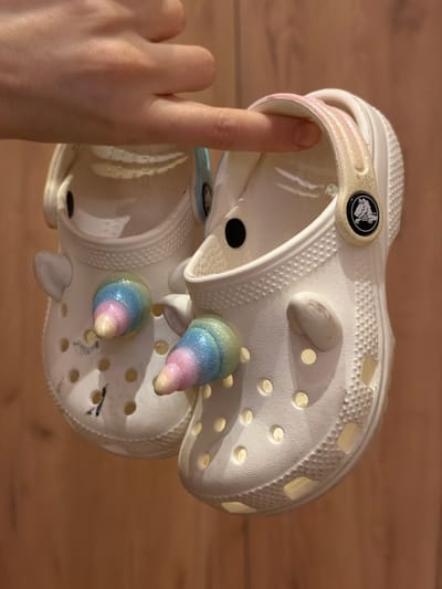 A human hand, characterized by a light skin tone, suspends two white Crocs brand clogs against a light brown wooden panel background in Rzhavki 2, Russia. An index or middle finger passes through the glittery light pink and gold strap of the right clog, while a thumb rests against the strap of the left clog. Both clogs are adorned with two iridescent, glitter-coated unicorn horn Jibbitz charms, featuring a blue, pink, and yellow gradient, along with small white ear-shaped charms. The right clog displays the black and white Crocs alligator logo on its strap button. The left clog exhibits dark scuffing or marks on its upper front surface. The background consists of vertically oriented wooden planks.