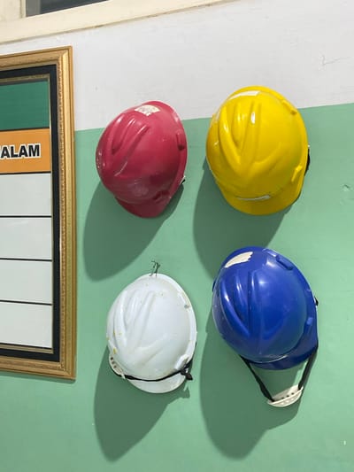 A collection of four hard hats is displayed on a wall, each suspended by a strap. The hard hats are in four different colors: yellow, blue, red, and white. They are arranged in a 2x2 grid. The yellow and blue hard hats are in the top row, with the yellow on the left and the blue on the right. The red and white hard hats are in the bottom row, with the red on the left and the white on the right. The wall is painted in two different colors, white on the left and a light green on the right. A framed picture hangs horizontally below the hard hats. The picture frame is gold with decorative detailing. Below the frame, a portion of a chart or poster is visible with vertical black lines and some text. The word "ALAM" is visible in large, bold, orange lettering on a darker background, part of the poster. The lighting suggests it could be daytime, with shadows cast by the hard hats indicating a light source from the left. The overall scene appears to be indoors, possibly a storage area or workshop, in Margoyoso, Indonesia. There are no people visible in the image.